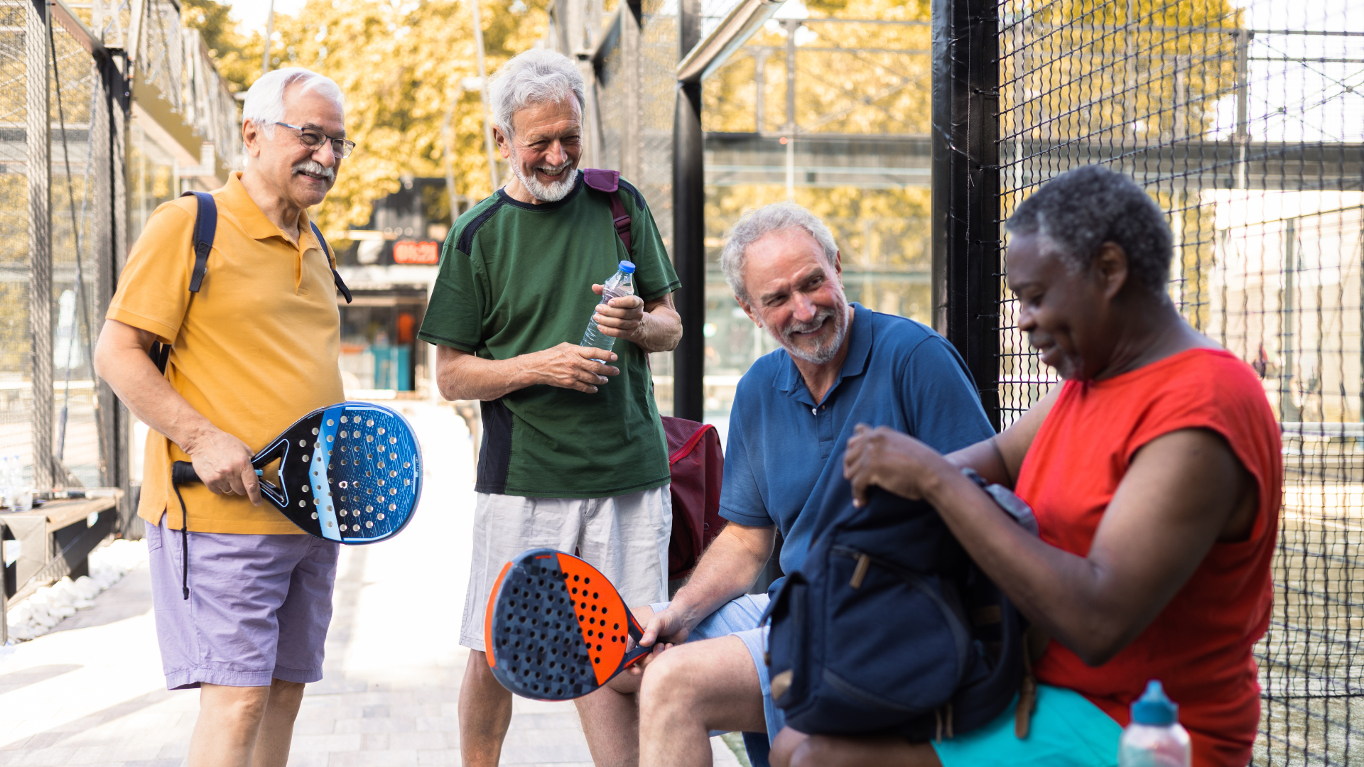 A group of four older people are gathered outside the local tennis court with pickleball rackets and equipment.