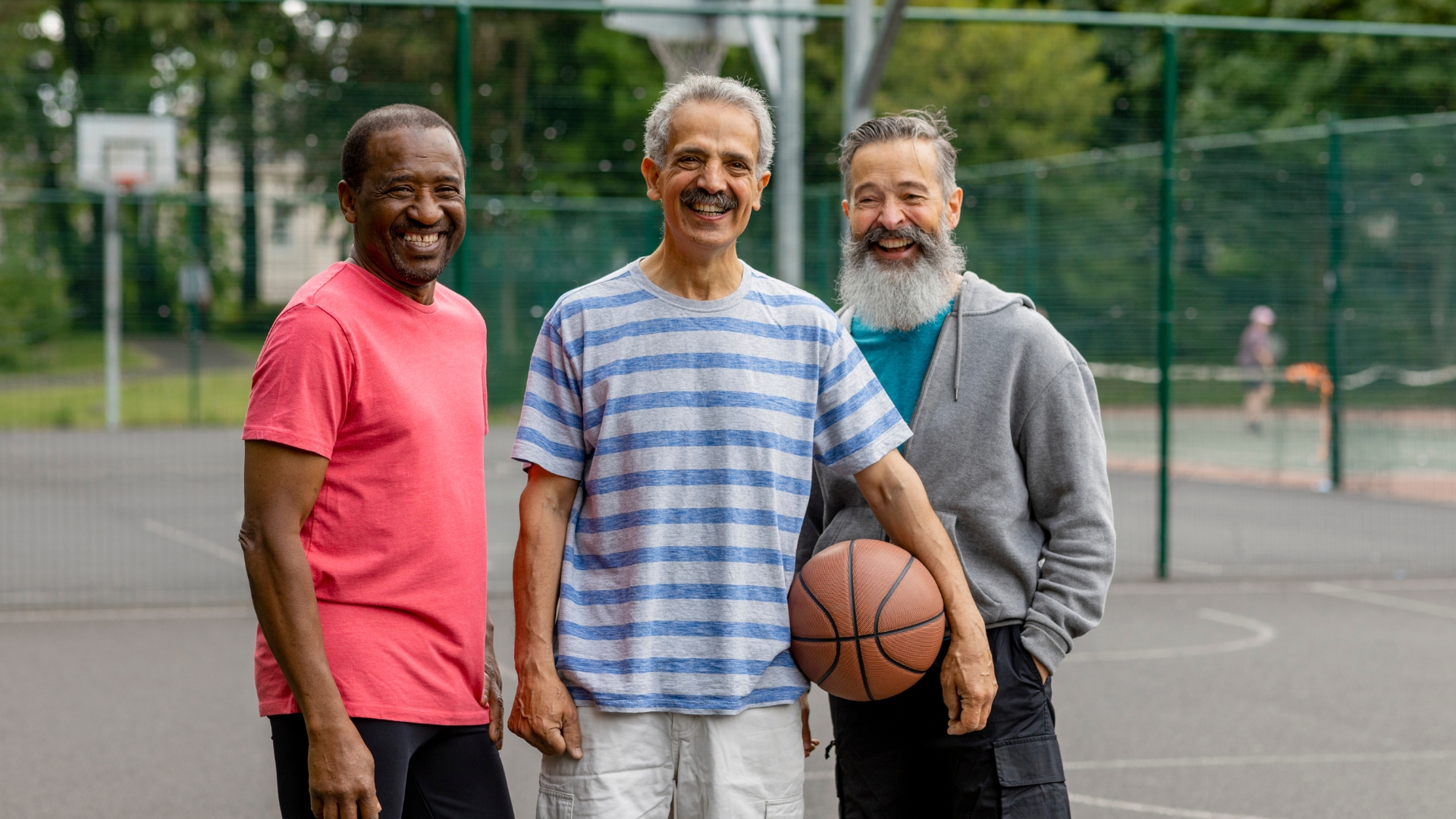Three older men are standing on the basketball court. The one in the middle is holding a basketball.