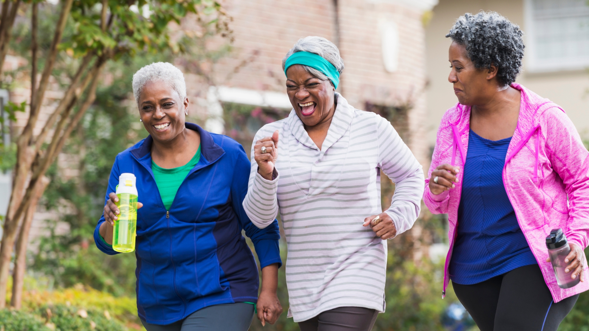 Three older women jog. The two on the sides are holding waterbottles and wearing jackets. The one in the middle has a joyous expression on her face